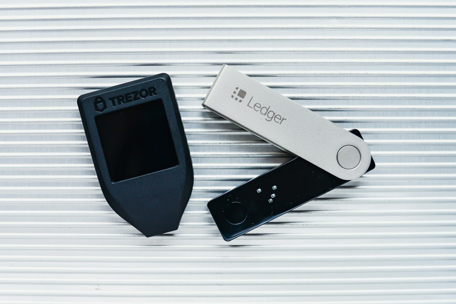 A Trezor Model T beside a Ledger hardware wallet on a metal desk